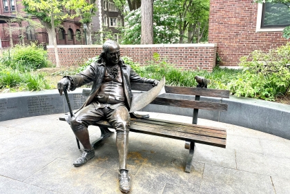 Bronze statue of Benjmain Franklin sitting on a bench reading the Pennsylvania Gazette