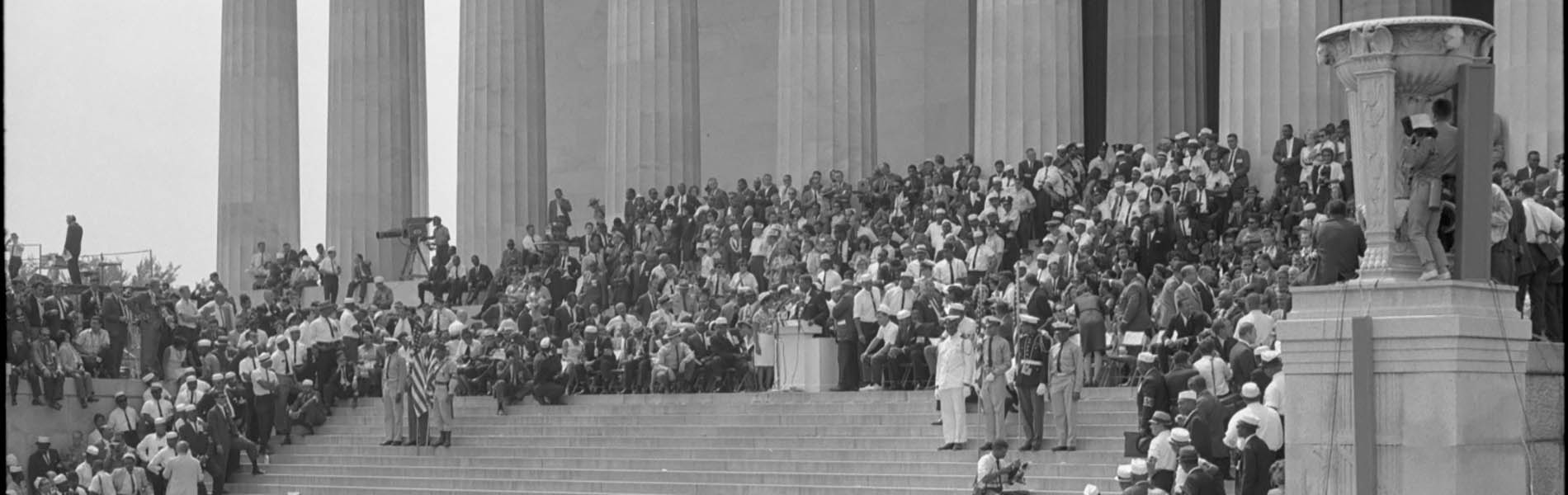 Black and white photo of crowd gathered for public address on steps of neoclassical building