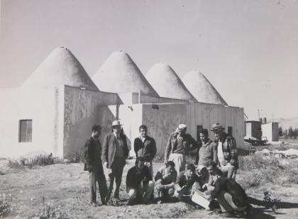 B&amp;W photo of people standing outside of building with several conic structures for a roof