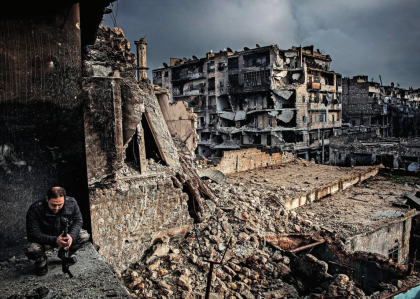 Man squats in foreground of collapsing buildings in Aleppo