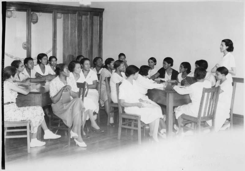 B&W photo of group of women having meeting around several tables