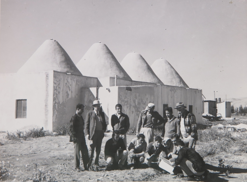 B&W photo of people standing outside of building with several conic structures for a roof