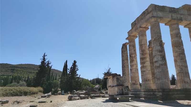 Sanctuary of Zeus at Ancient Nemea, view capturing the impression of the Temple of Zeus and sacred grove relation in ancient time. At the foreground is the restored temple’s east facade with ramp access and the remains of the long altar, at the background vegetation that includes cypress trees. The position of the trees that are seen today is not exactly that of antiquity, photo by Antonios Thodis. «The copyright of the depicted monuments belongs to the Hellenic Ministry of Culture and Sports (ν.3028/2002)»