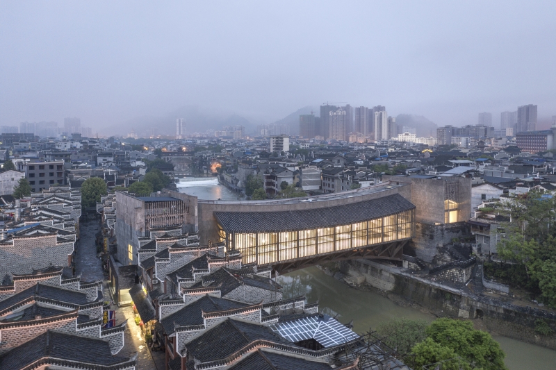 A lantern-like bridge in concrete and wood spans a small river in a dense urban setting