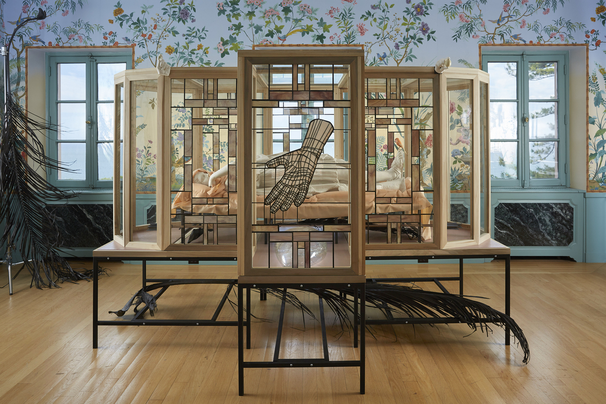 installation view of glass box with body inside, stained glass hand obstructing the view, in an ornate room