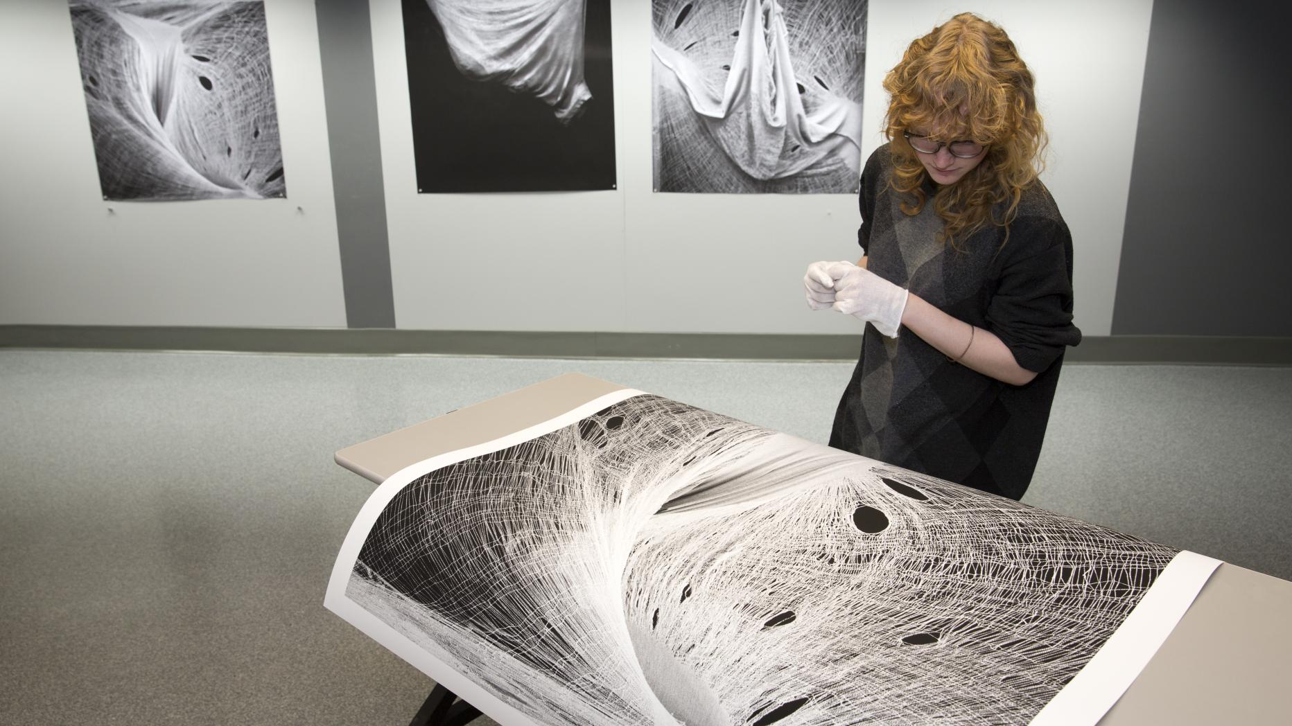 Woman looking down at a table with a large photo of her textile art with three textile photos on the background wall