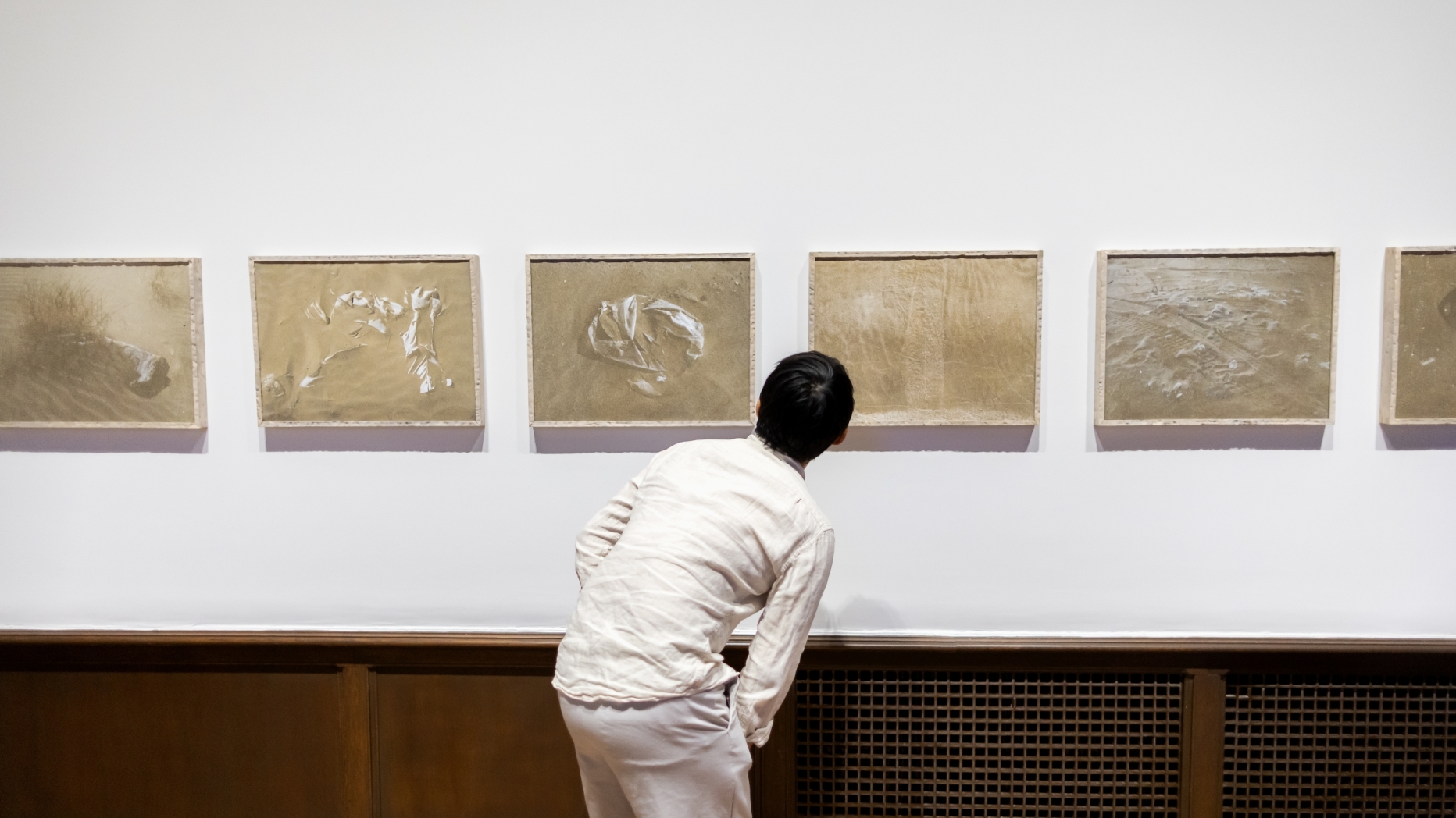 A person looks closeup at a row of works on gallery wall