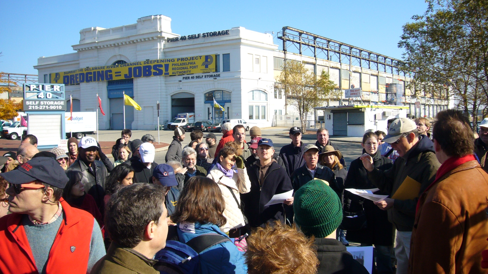 Group participating in the Central Delaware riverfront planning process gathers on a street corner and listens to an event organizer read from a document on a clipboard.