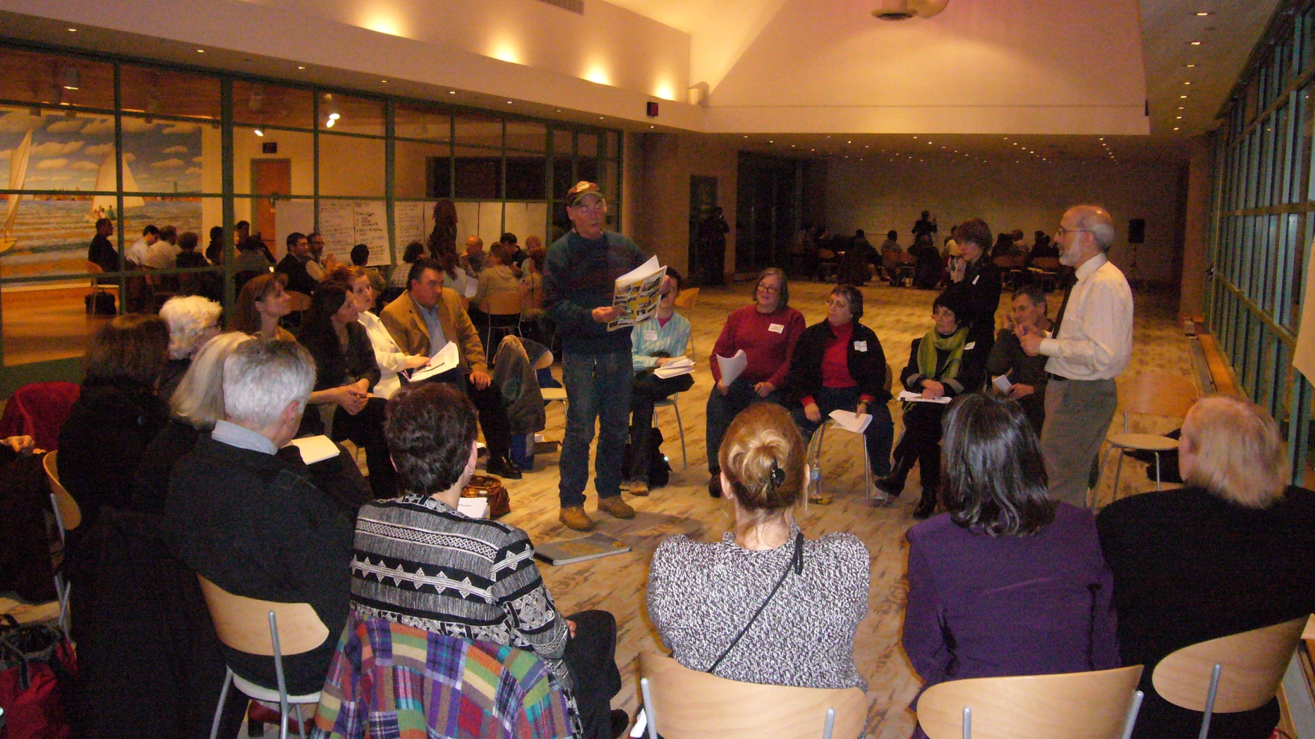 Project participants sit in a circle of chairs. A man stands in the middle and speaks while holding a large book.