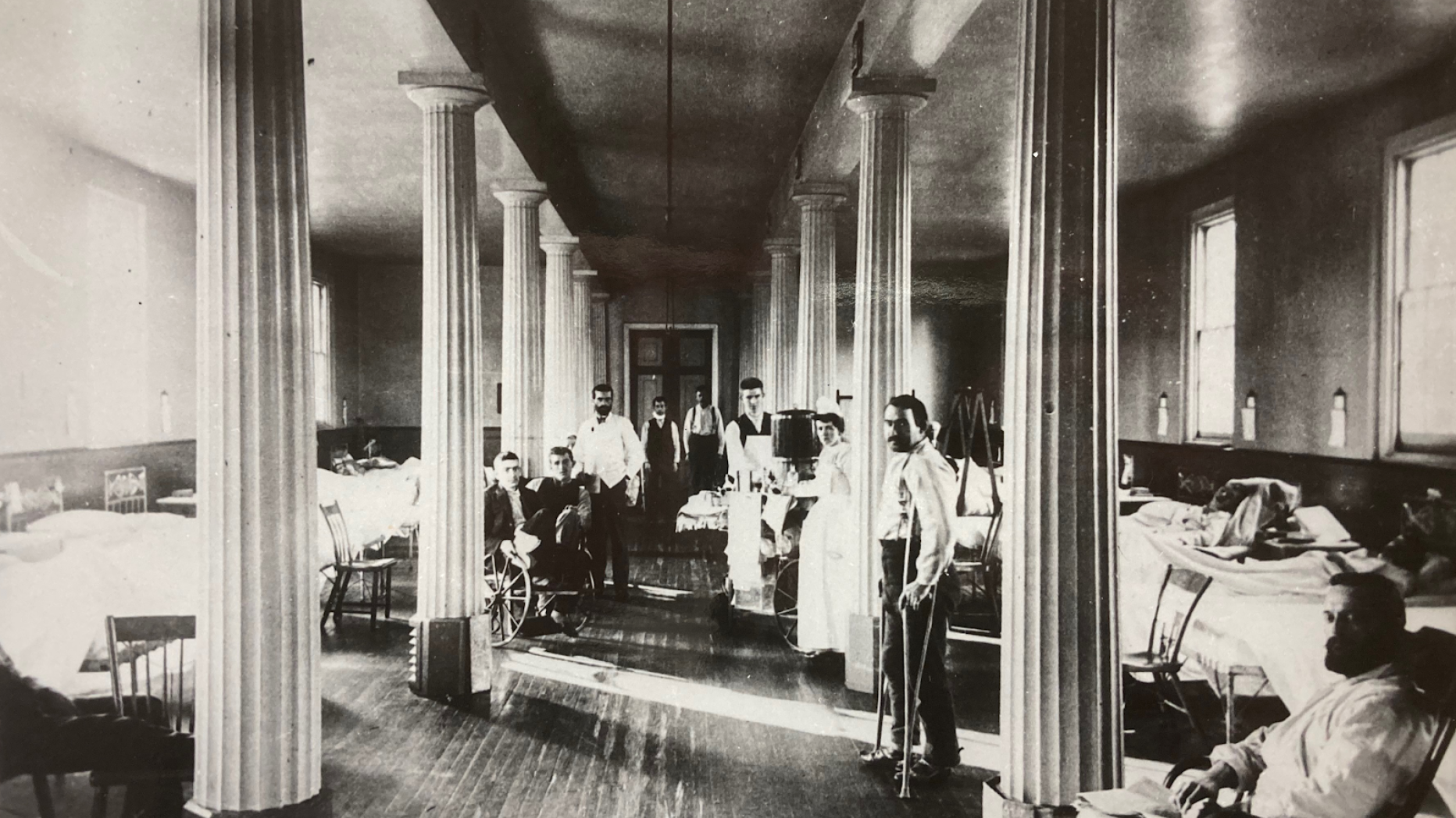Old black and white interior photo of hospital patients in a grand looking door lined with columns down the center.