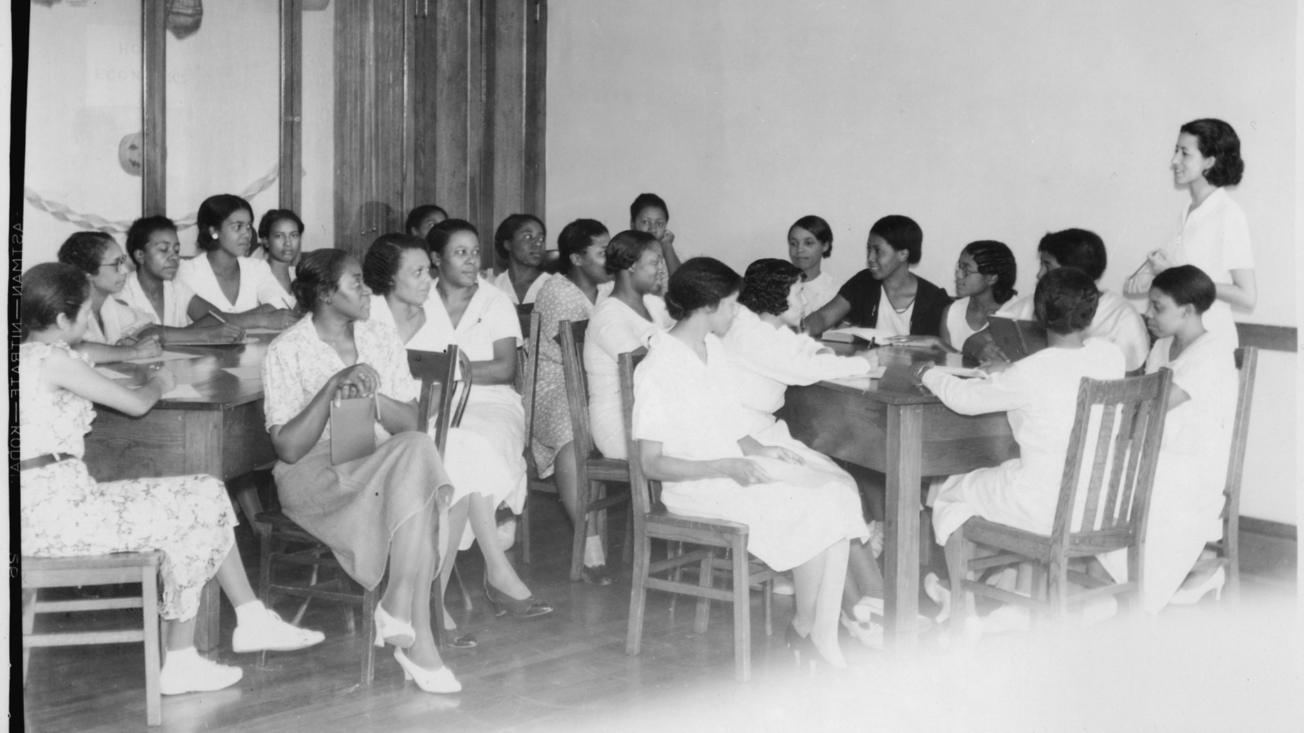 B&W photo of group of women having meeting around several tables