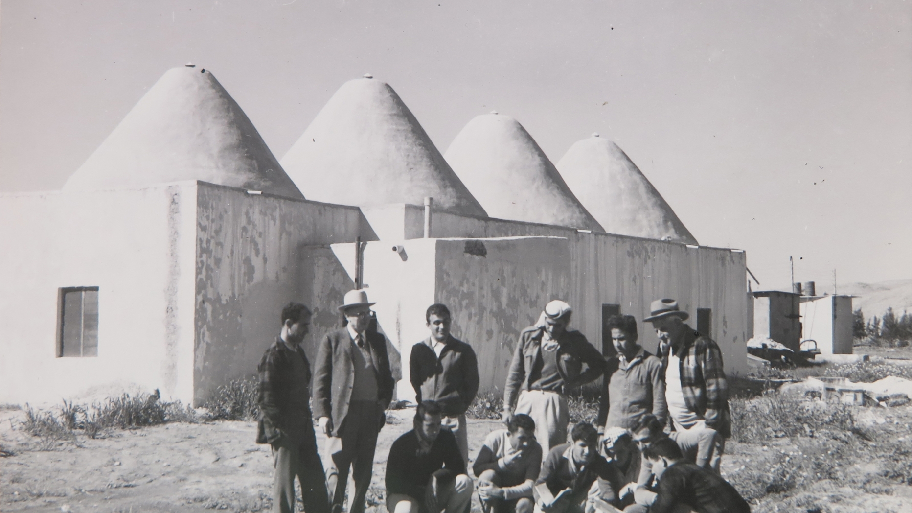 B&W photo of people standing outside of building with several conic structures for a roof