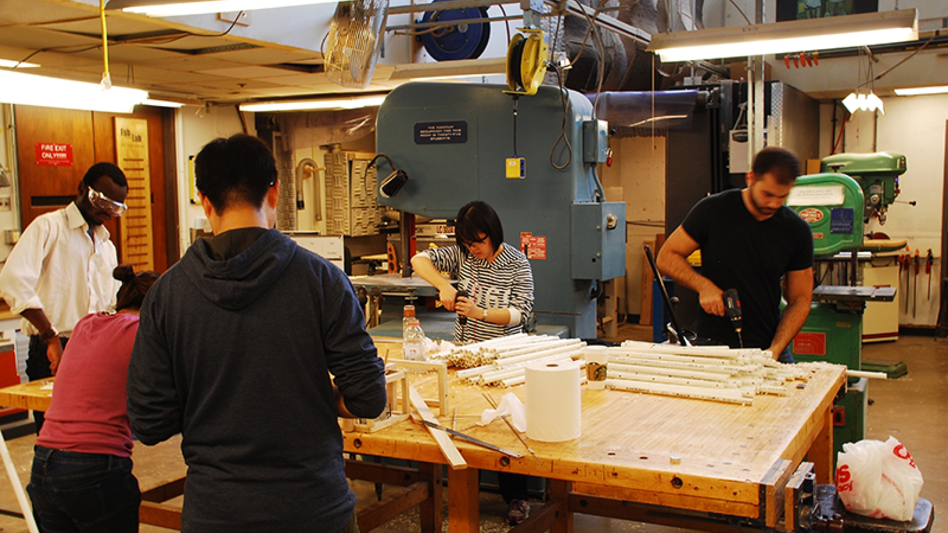 Group of students working at table with pbc plumbing and band saw in background