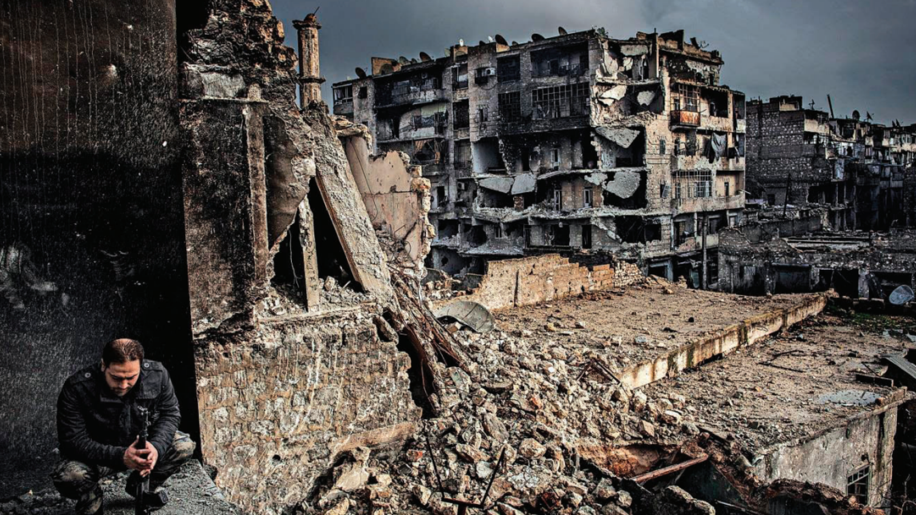 Man squats in foreground of collapsing buildings in Aleppo