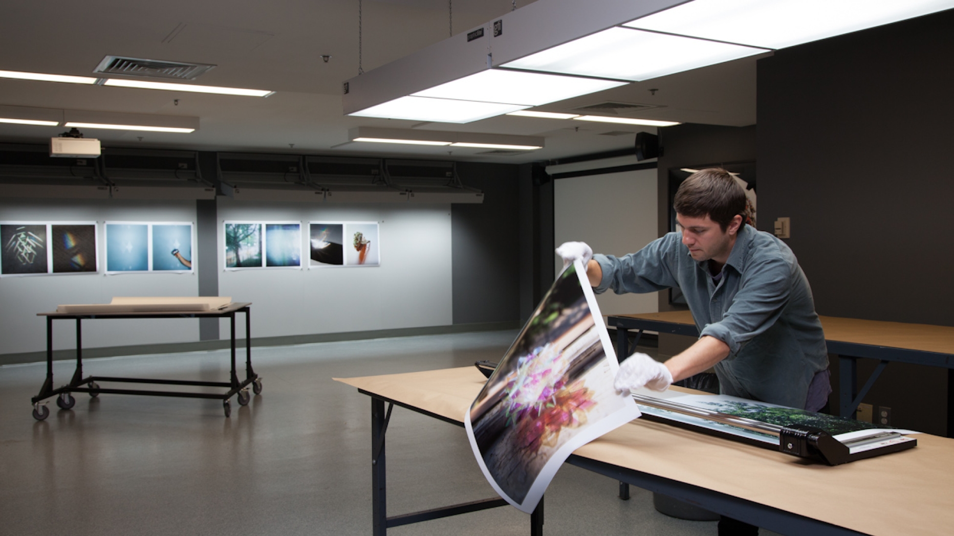 Student prepares to trim a large photograph in a softly lit studio. Large prints of photographs hang on the wall in the back