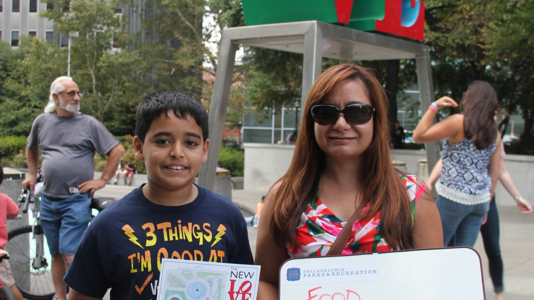 Woman and child standing in front of LOVE statue with a sign saying their suggestions for the improvement of the park ("Food, flowers, trees!").