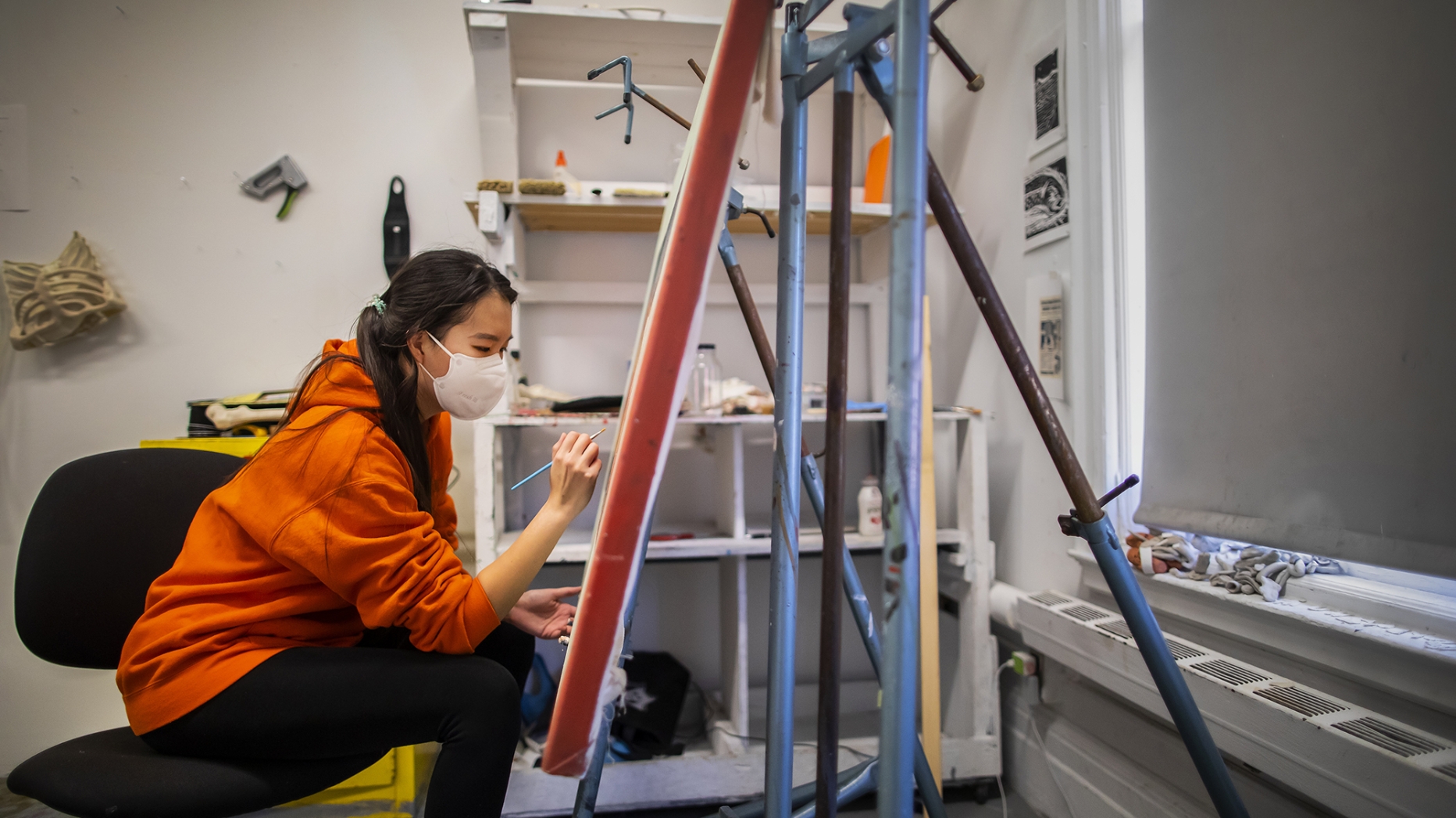 student in orange hoodie crouched and painting at an easel 