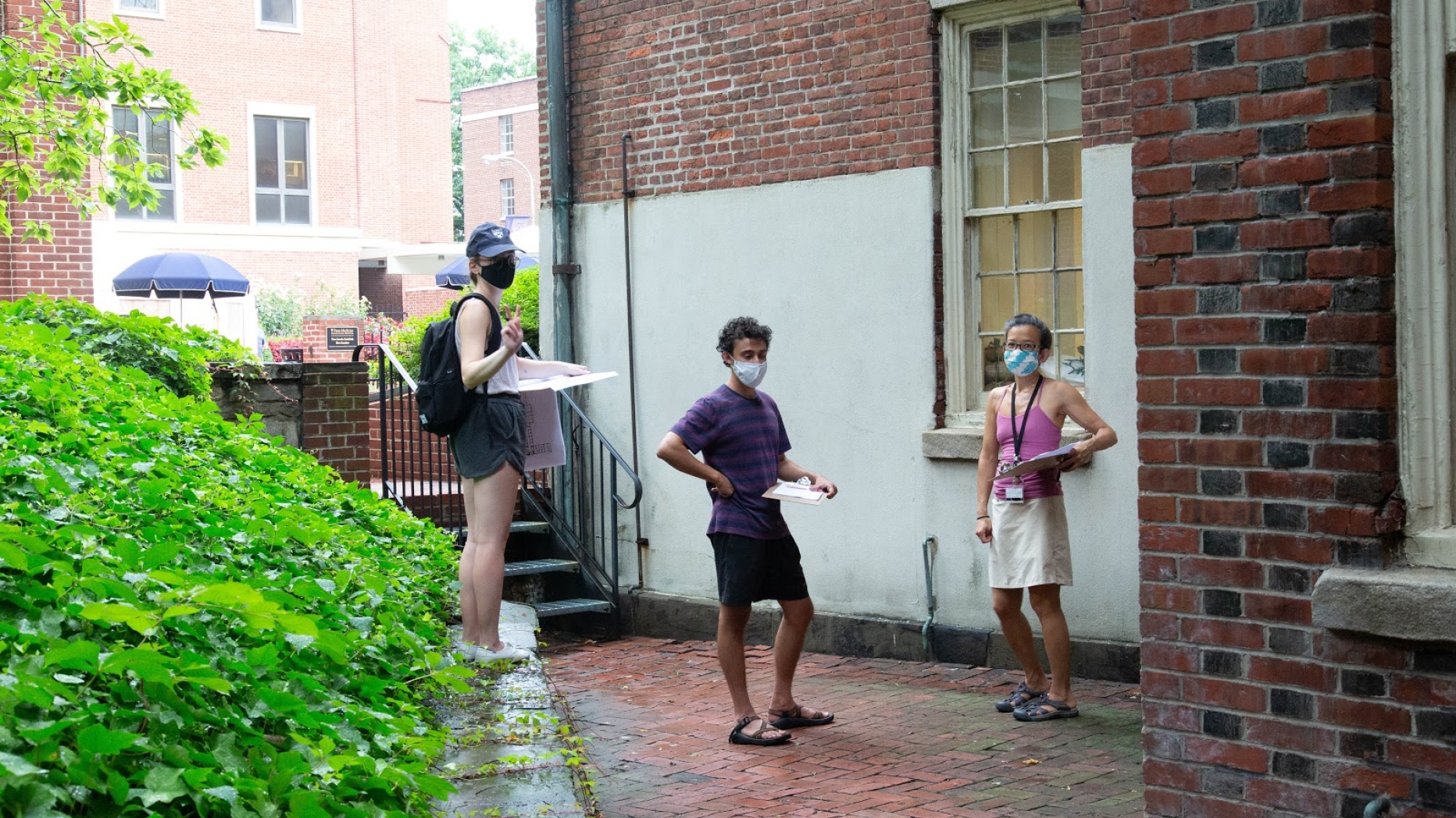 Three researchers with clipboards, masked and distanced, face the camera posed against a brick building