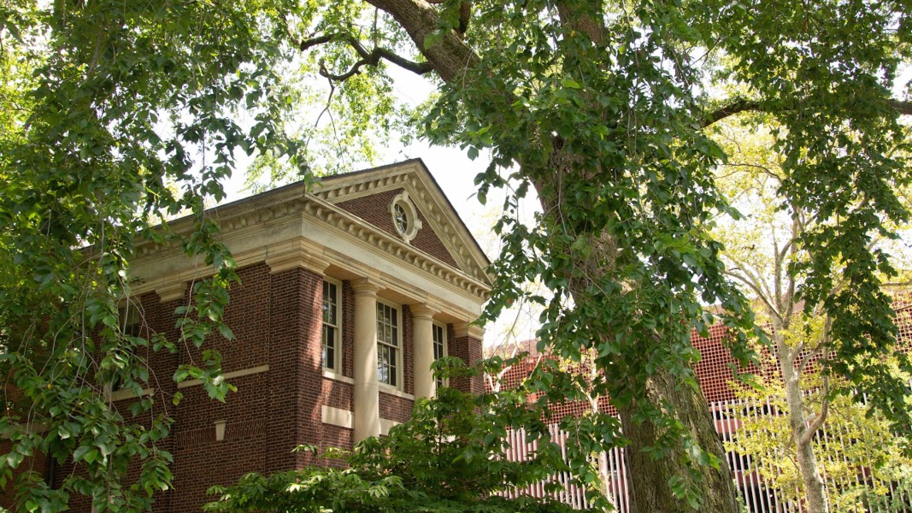 Lush landscaping surrounds a brick building with doric columns. A large sweeping free is in the foreground. 