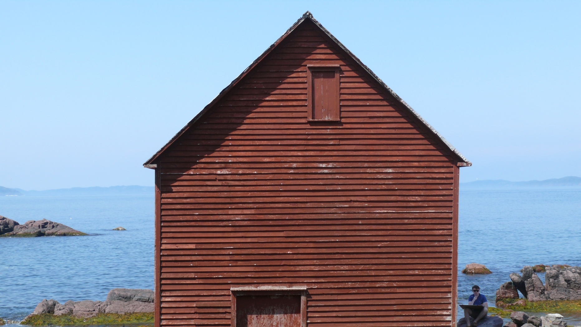 front gabled building on shoreline with rust-red paint on wood paneled facade