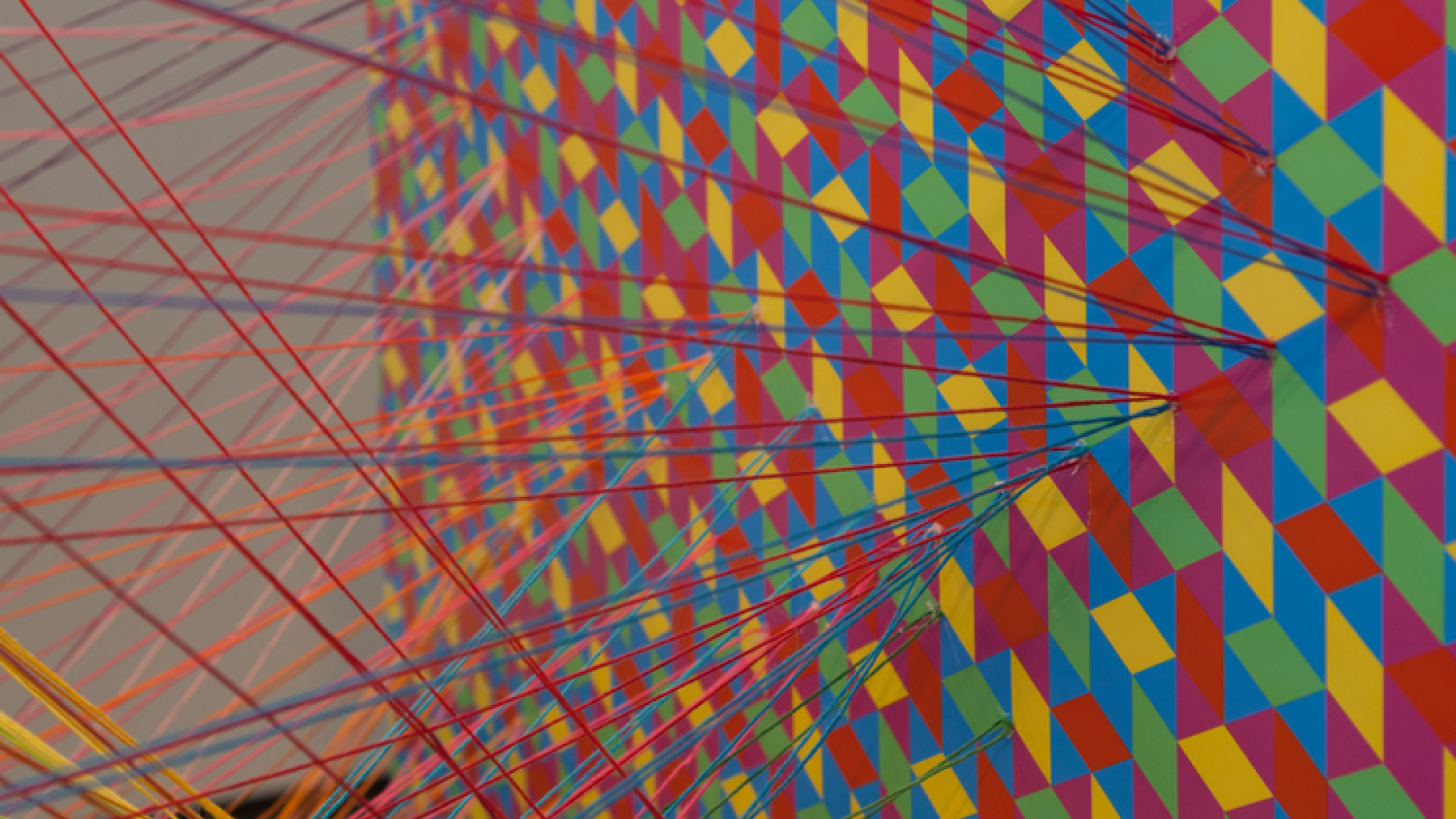 Art piece where multicolored yarn is attached to a board with colorful pattern. Yarn is suspended in space in a criss cross pattern similar to that on the board.