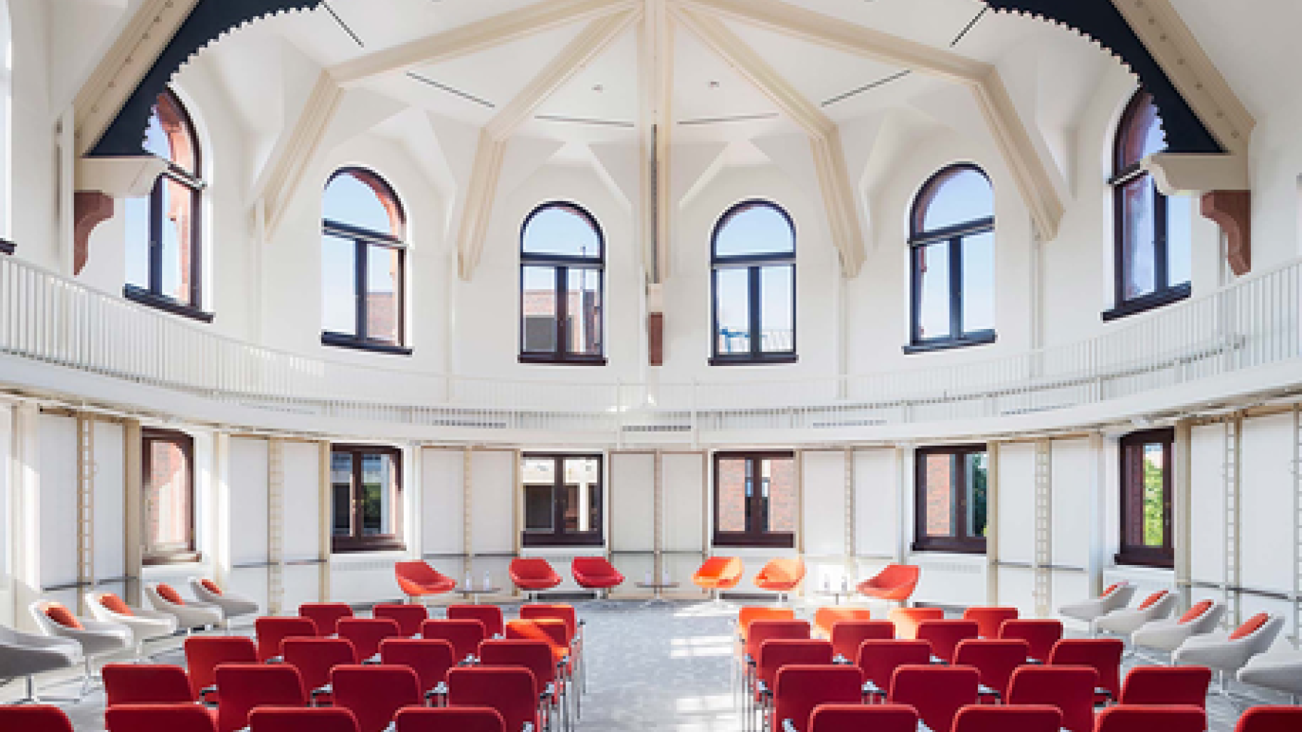 Spacious white room with lots of windows. Rows of red chairs face the front of the room where a number of chairs are set up