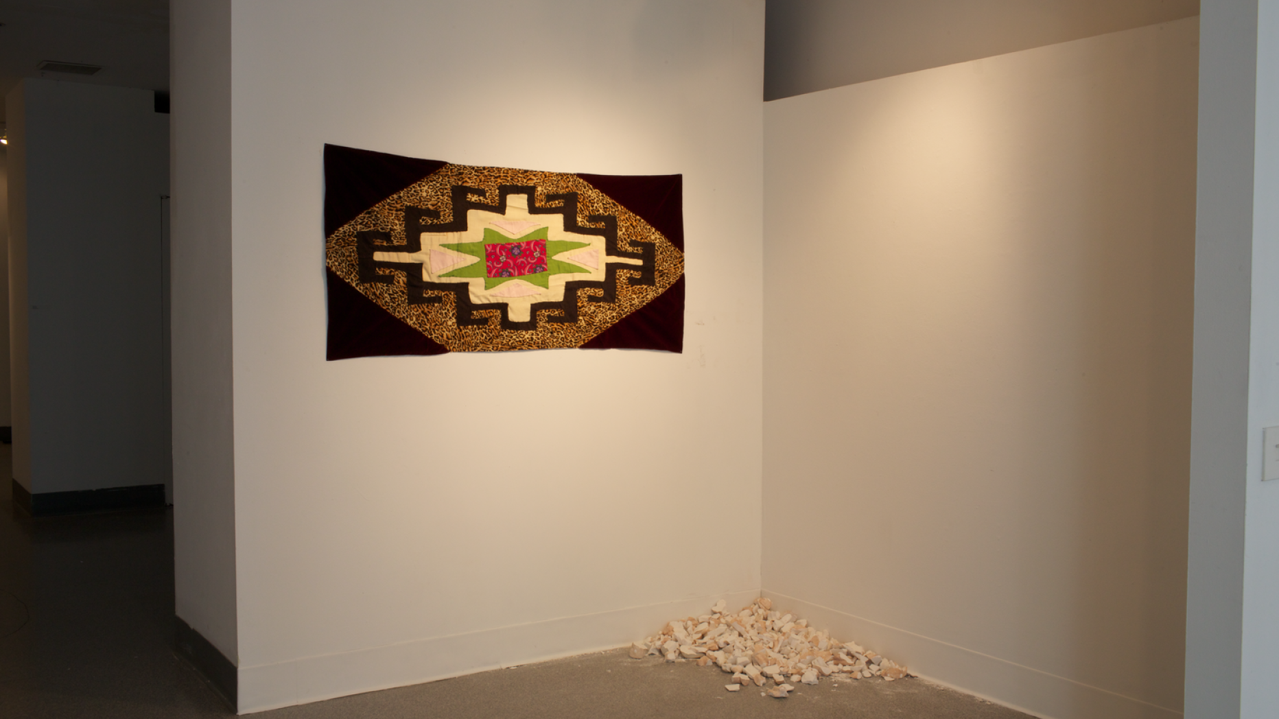 A gallery space with a patterned quilt on the wall and a pile of crumbled material in the corner.
