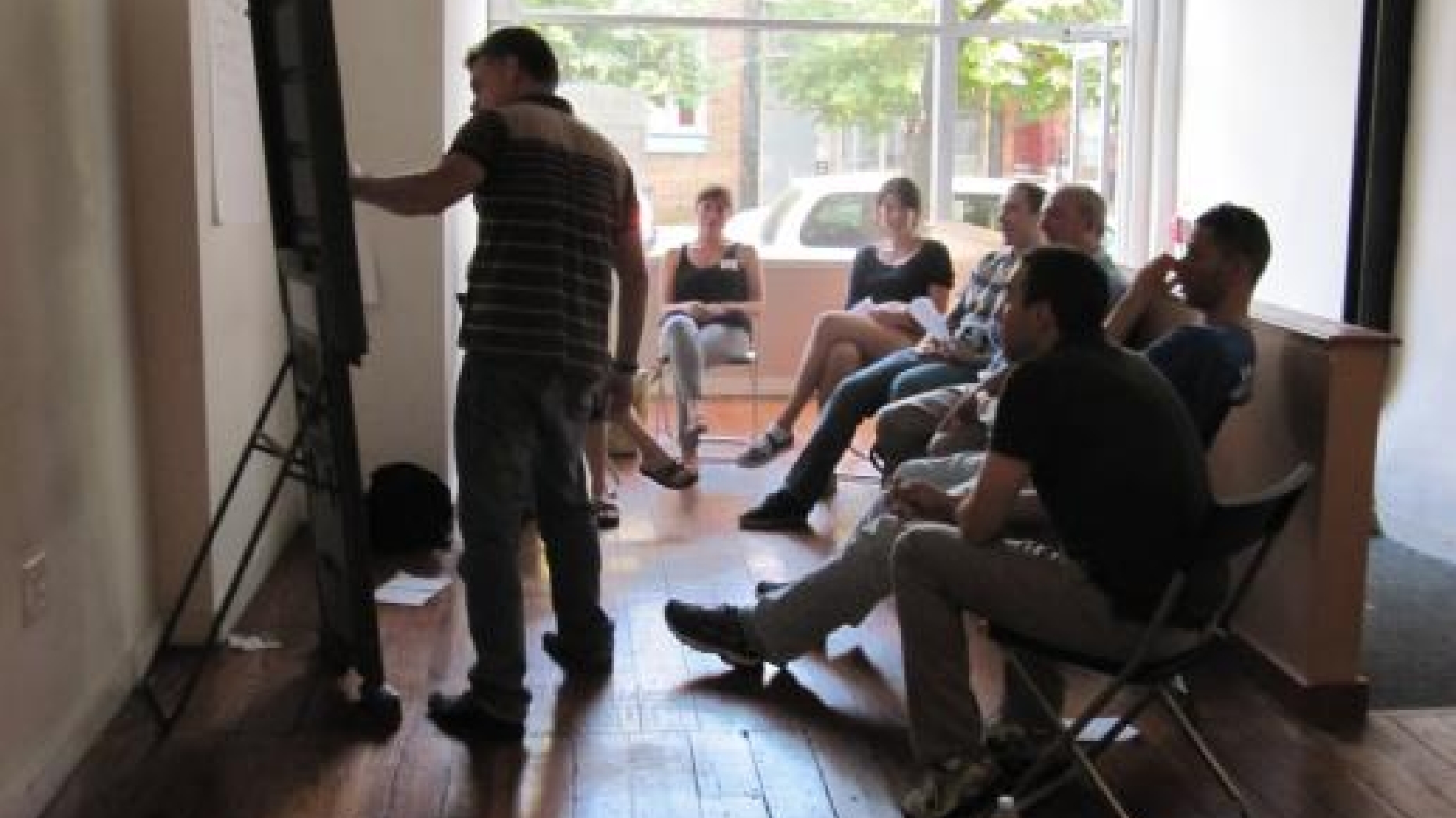 Project meeting. Group sits in a semicircle of chairs looking at a man talking about something written on a drawing pad.