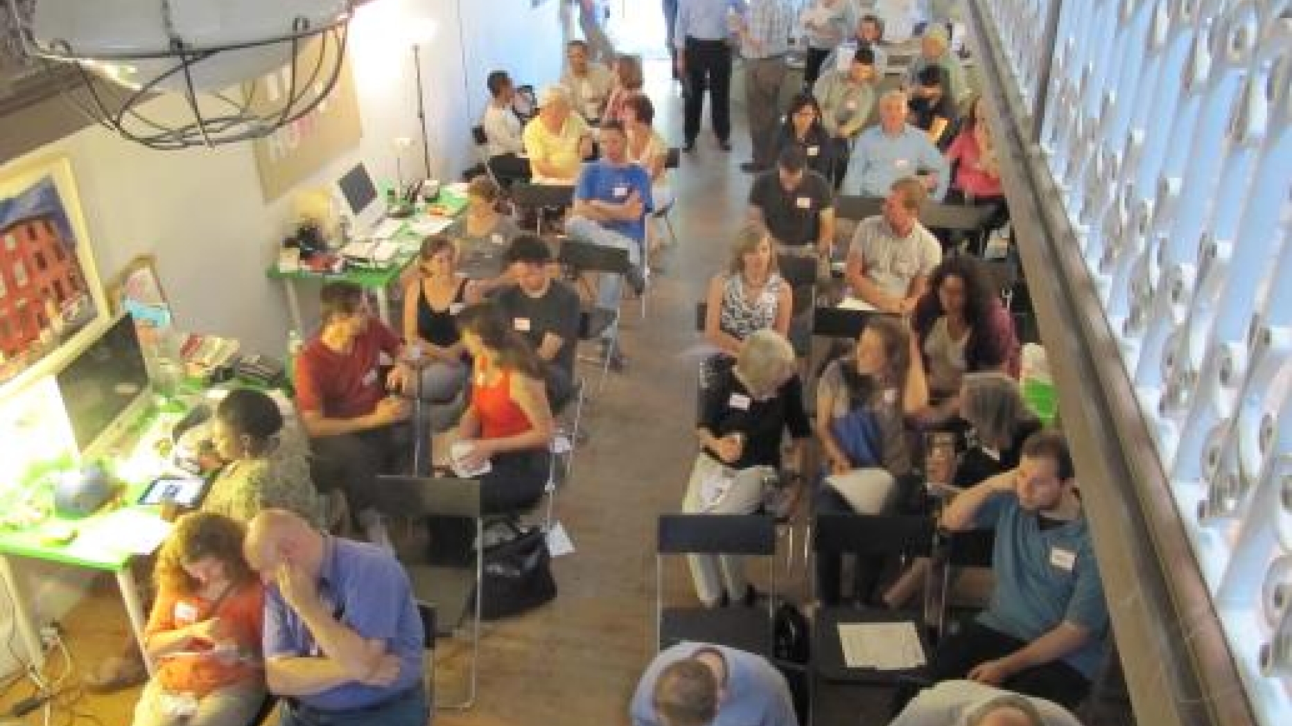 Large meeting of the project. Many people in chairs in a long room also containing a few computer work spaces.