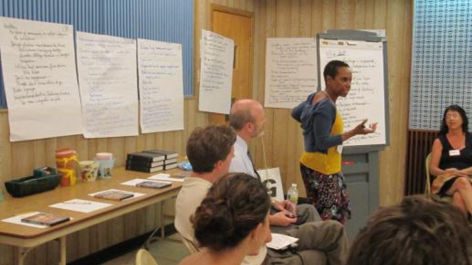 Group meets in a circle. Many ideas are posted on the wall behind them. One person stands by a drawing board and discusses what is written on it.