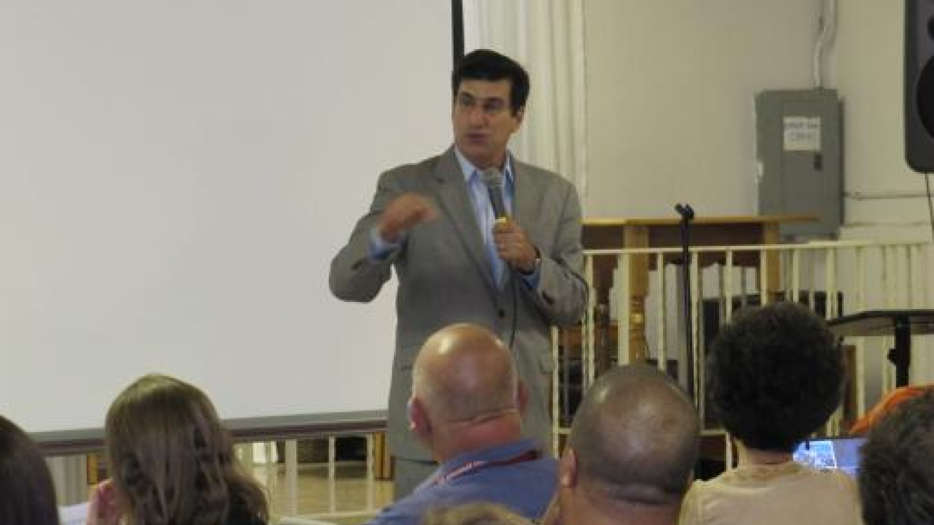 Man stands in from of white board with a microphone speaking to an audience seated before him.