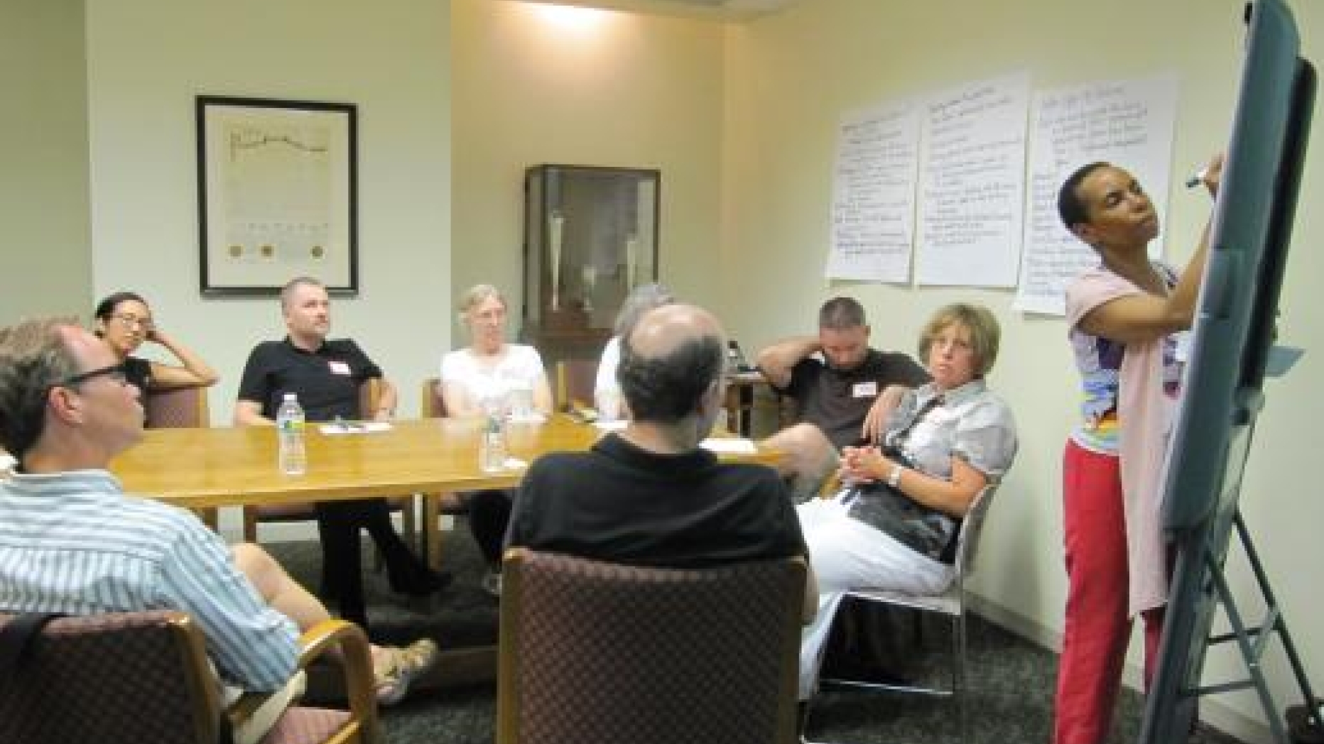 Group meets around a table with project ideas posted on the wall behind them. One woman is standing and writing further ideas on a drawing board.