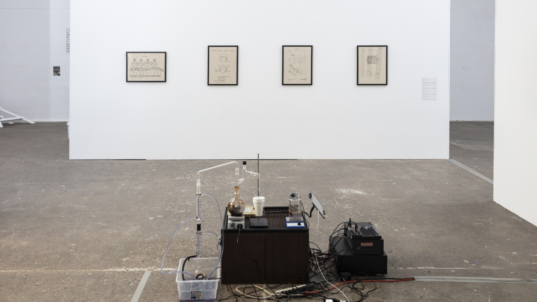 Gallery space with multimedia installation involving chemistry equipment, electronics and blueprints on wall in background