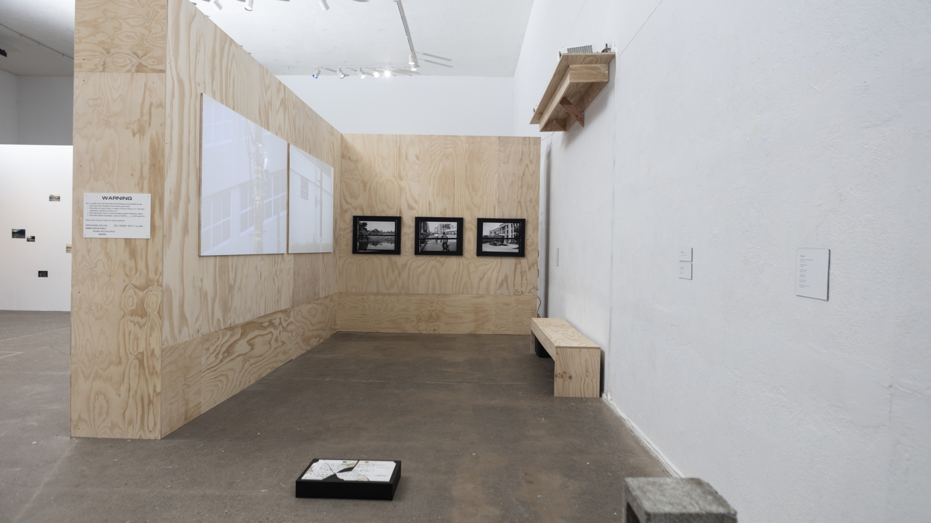 Gallery space with alcove made of plywood. Photographs are hung on wall and there is a bench to sit and view them