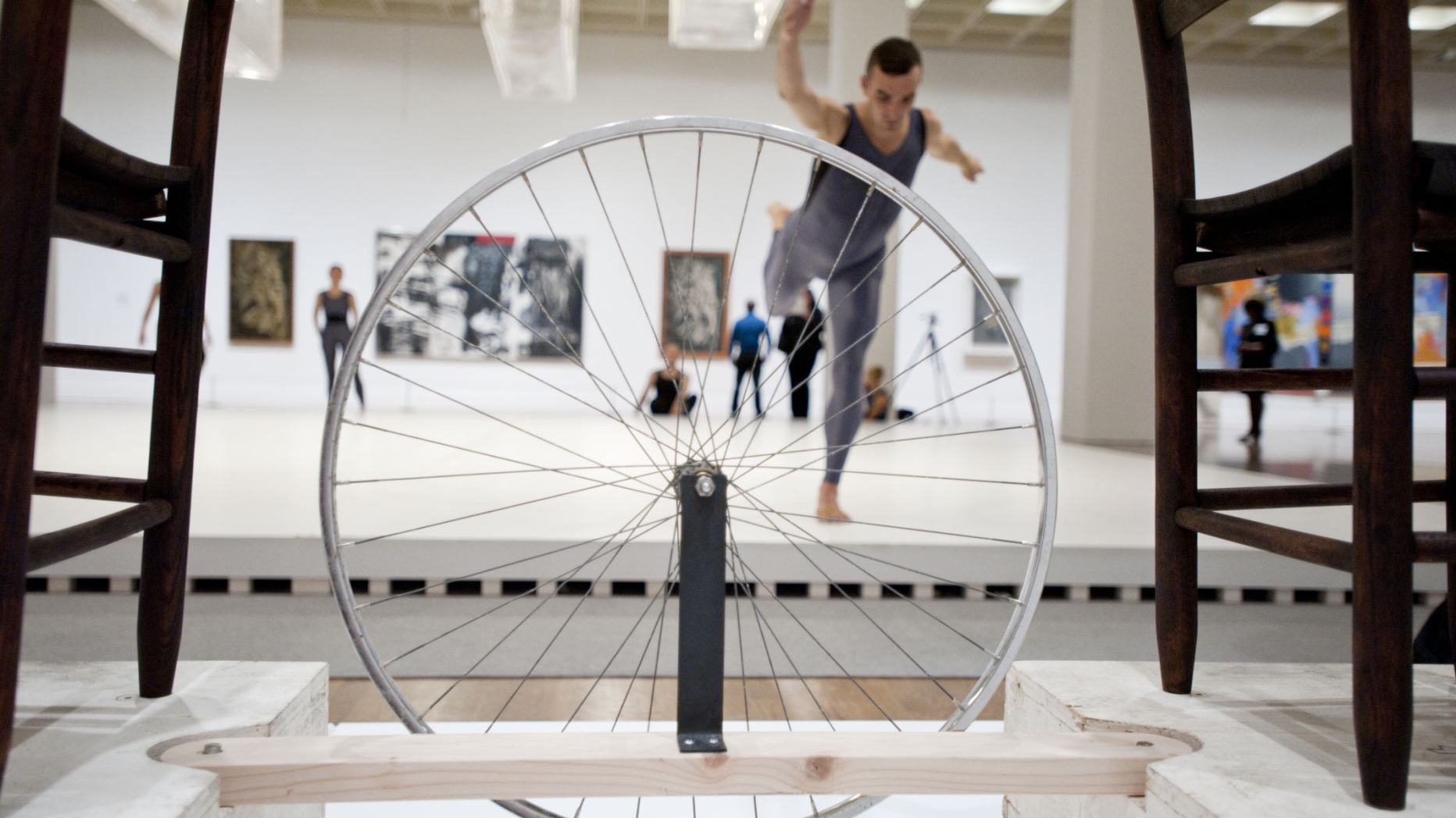 Foreground: sculpture with bicycle wheel suspended between two chairs. Background, performers dancing