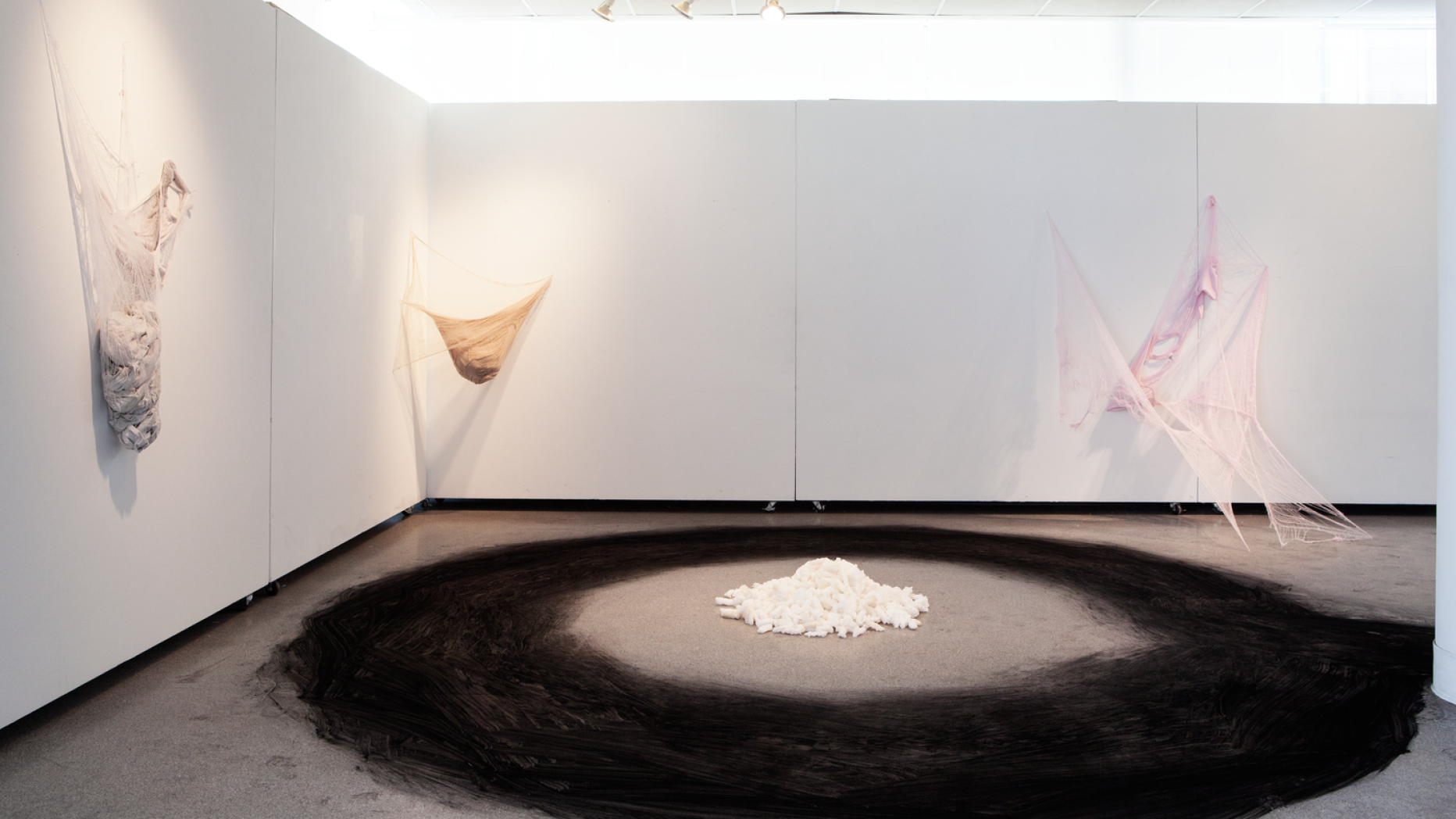 Gallery space with three pieces utilizing yarn and fiber on the wall and a large piece on the middle of the room composed of a large black ring painted on the floor with a small pile of white rocks in the center.