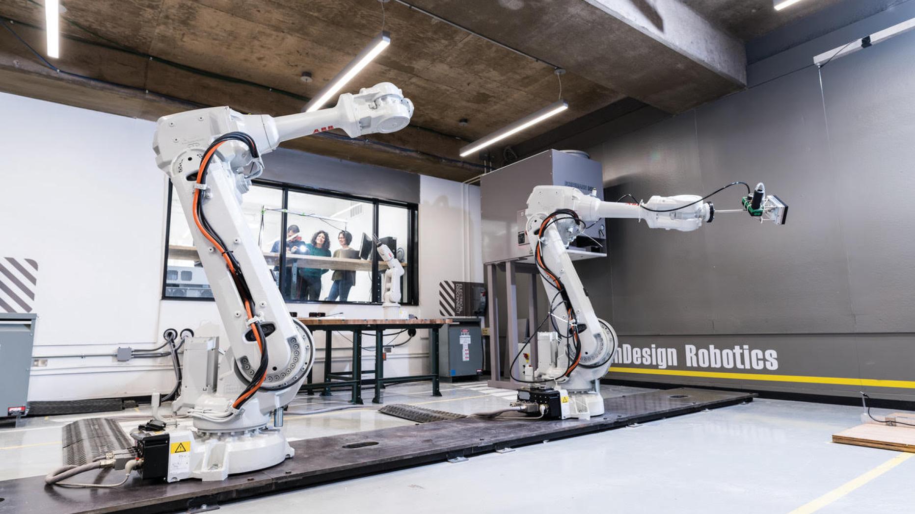 Two large robotic arms in a studio with students viewing them through a window to an adjacent room