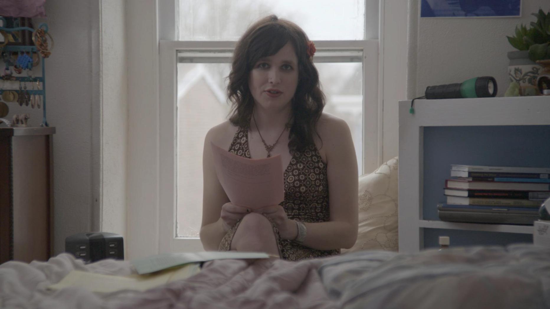 Woman looking at camera holding a piece of paper with a bed in front of her