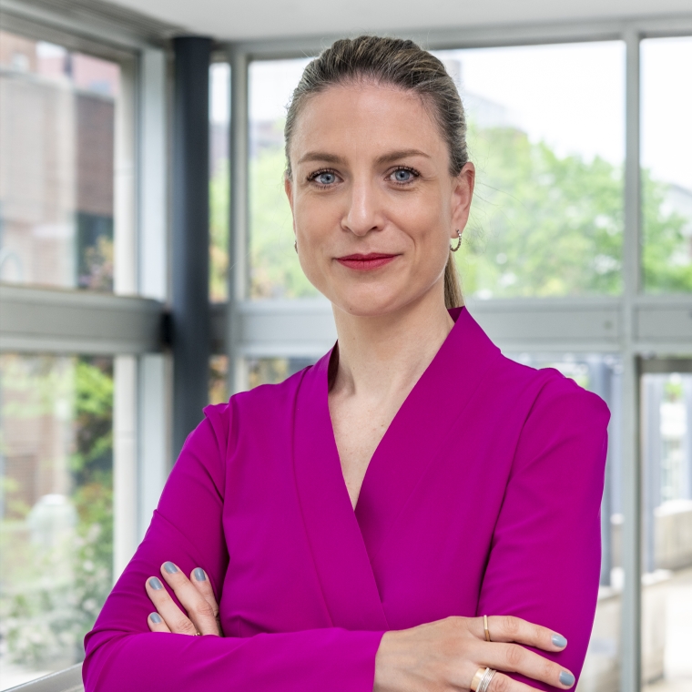 headshot of Zoe Ryan, arms crossed across her bright pink blazer and hair pulled back, in a greatly lit all window room.