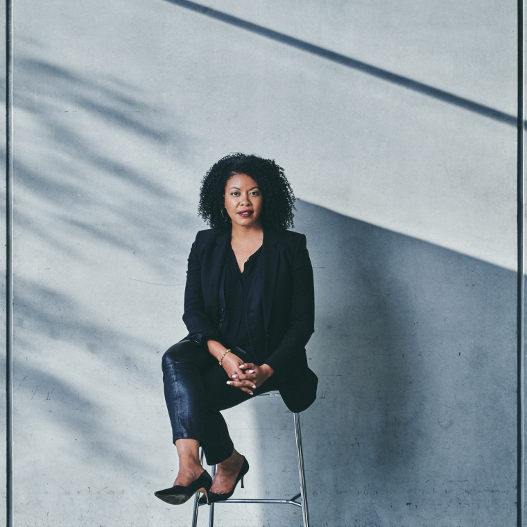 Photograph portrait of Adrienne Edwards sitting on a stool against a light-colored wall