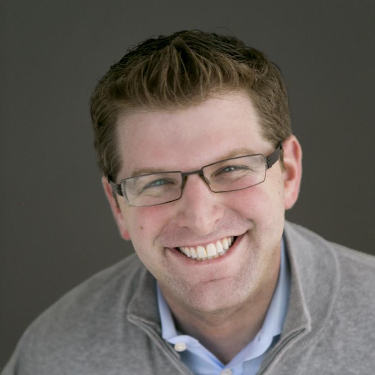 Rachlin faces the camera, smiling, wearing glasses and set against dark gray background