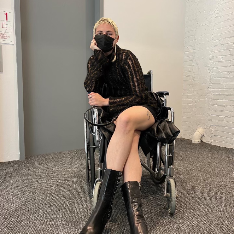 Photograph of Mae Eskenazi in front of an elevator in a wheelchair wearing black face mask and black boots