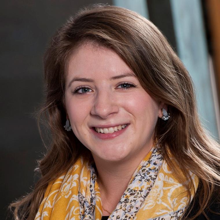 Lake smiles at the camera, looks straight ahead, has dark blonde shoulder length hair and wears a colorful yellow and blue scarf