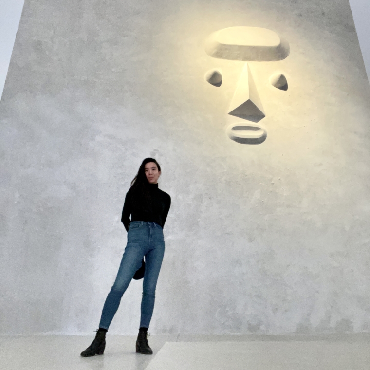 Photograph of Xin Wang standing in front of a light gray concrete or stone wall with a low-relief sculpture of an abstract face on the wall above.