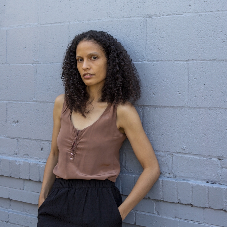 Vaughn leaning back on a blue painted brick wall, hands in her pockets