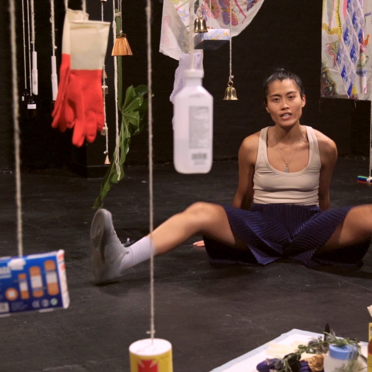 Performer sitting on ground in black box, with hanging objects in the foreground to background
