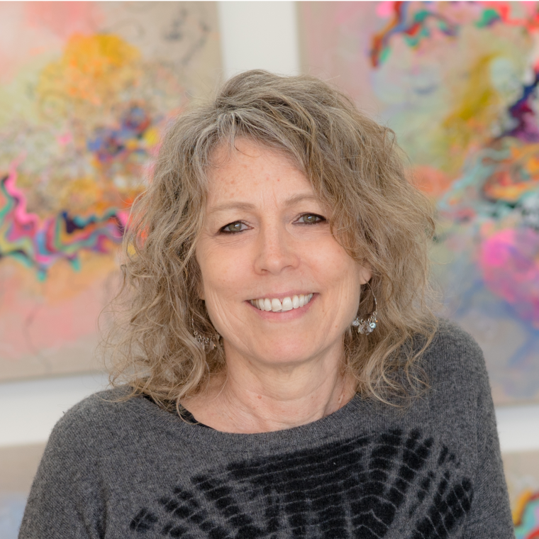 Photo portrait of Jackie Tileston smiling and sitting in front of two of her paintings