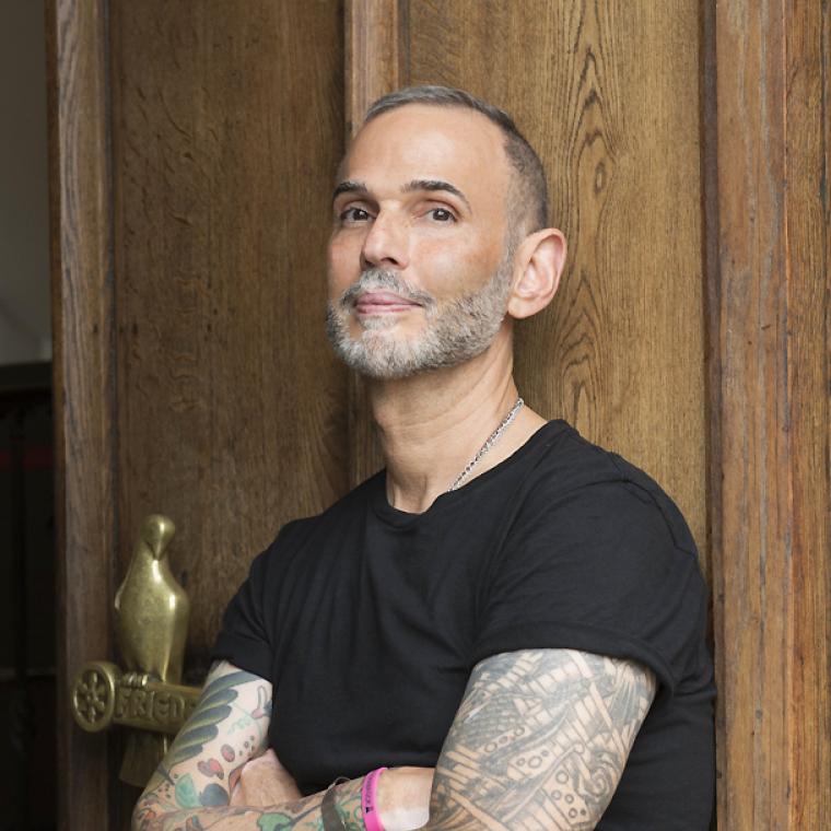 Headshot of Ernesto w heavily tattooed crossed arms, leaning against a wooden door frame