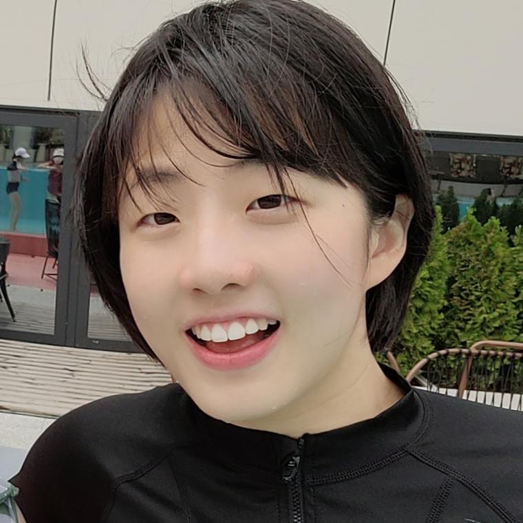 Photograph of Gayoung Lee smiling, wearing a black shirt, standing in front of a tile background