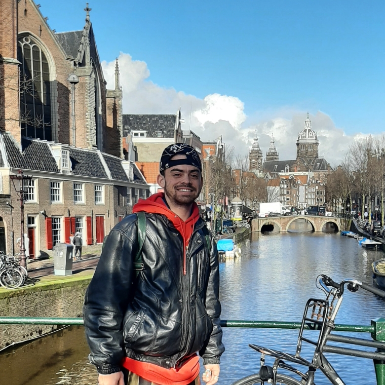 A photograph of Geo Wyex standing on a bridge in front of a river surrounded by historic houses